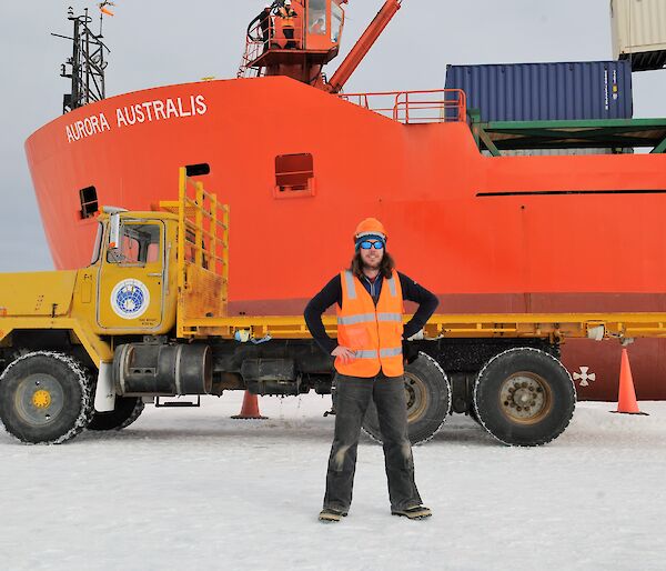 A man is standing on the sea ice in front of a yellow truck parked in front of a red icebreaker ship