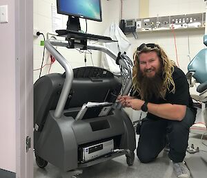 A bearded doctor is holding a screwdriver next to a grey x-ray machine in a dental surgery