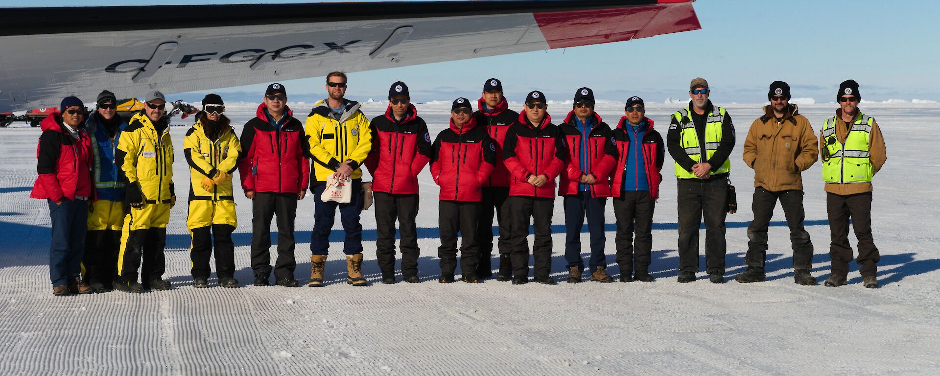 A group of people wearing yellow and red lined up under the wing of a plane