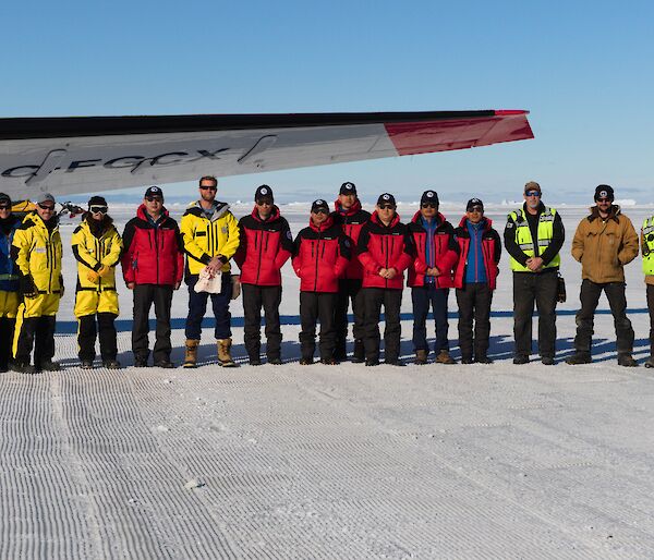 A group of people wearing yellow and red lined up under the wing of a plane