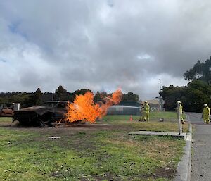 A car on fire with a big orange flame and three people standing nearby