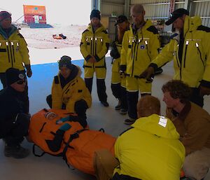 a group of people in yellow standing around a stretcher with a person wrapped in orange.