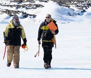 Two men walk across the blue sea ice while carrying equipment