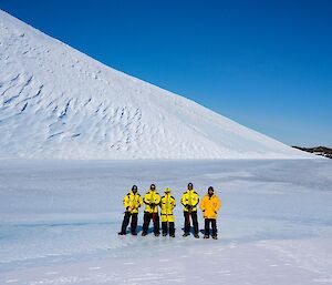 The group stand on a blue frozen lake surrounded by steep snow slopes