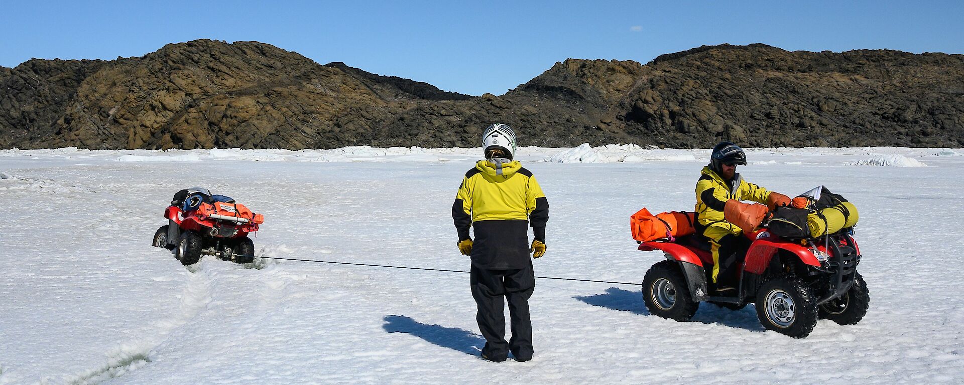A man stands beside another man on a quad bike as it is being pulled out of the ice