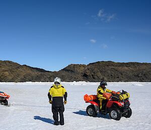 A man stands beside another man on a quad bike as it is being pulled out of the ice