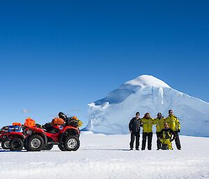 The field training group and their quad bikes pose for a group photo in front of a blue, triangular iceberg