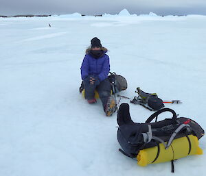 Flat, grey sea ice frames the shot as Scientist Louise rests on her field pack under a gloomy sky