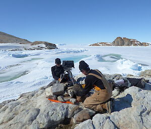 Two men install a camera at a sun-lit island location, surrounded by sea ice and other islands.