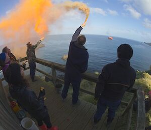 6 people stand on a wooden deck watching 2 orange flares with the Aurora Australis in the distance