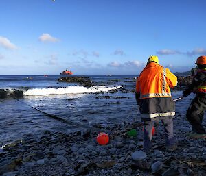 Two men in waterproof clothing are standing on a beach holding a fuel line, with the Aurora Australis in the background.