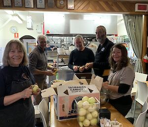 5 people stand around a table peeling potatoes and onions