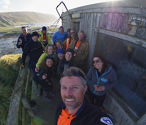 14 people stand on a wooden deck in front of a wooden shack and a green island