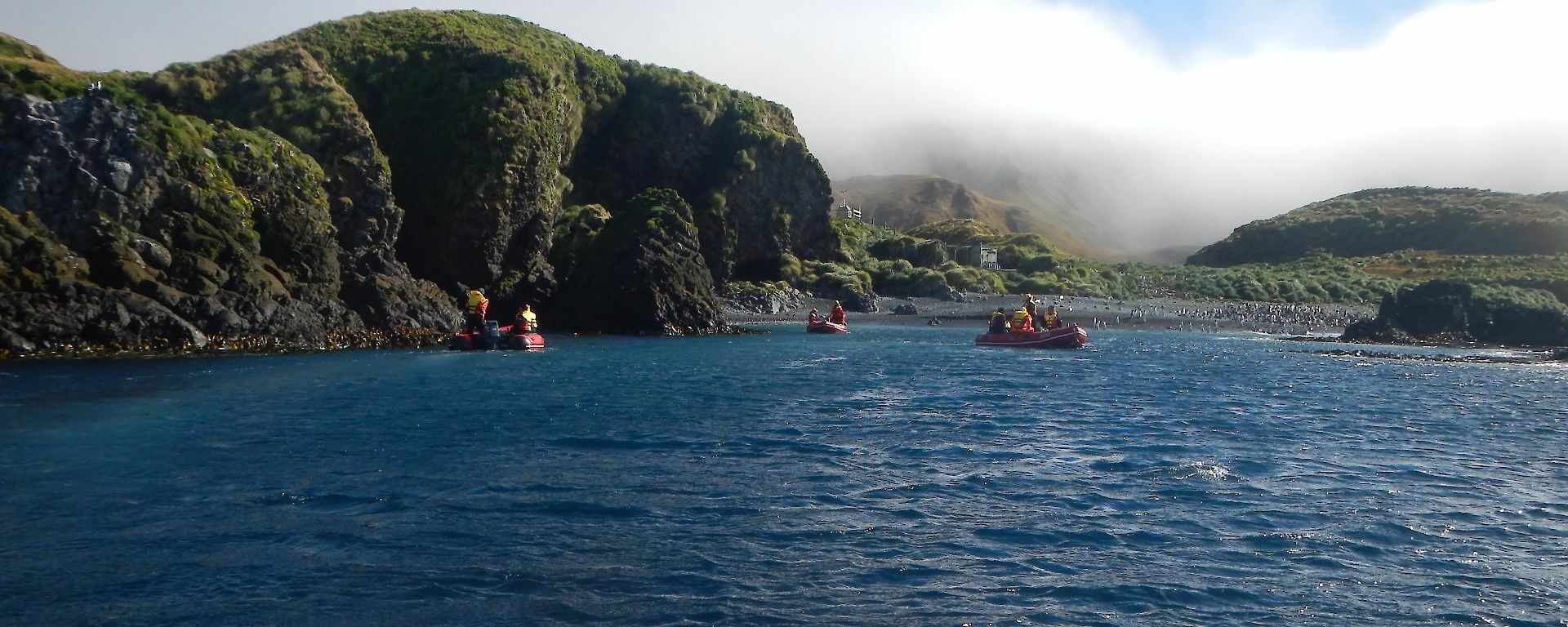 Three red inflatable boats are approaching a small bay with green outcrops in the background.