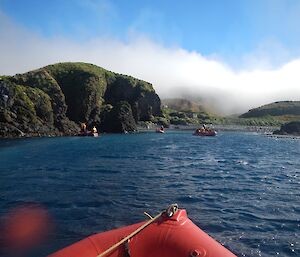 Three red inflatable boats are approaching a small bay with green outcrops in the background.