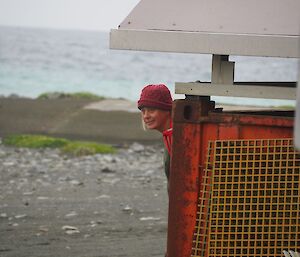 A woman is peeking around a shelter with a beach in the background