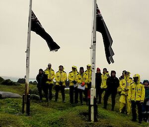 13 people stand in raincoats in front of flagpoles