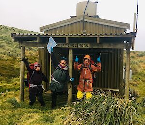 Three people stand in hiking gear in front of a wooden field hut