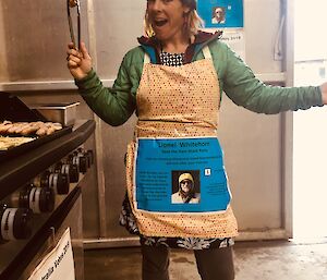 A woman stands in an apron holding a pair of tongs with a sausage