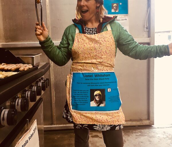 A woman stands in an apron holding a pair of tongs with a sausage