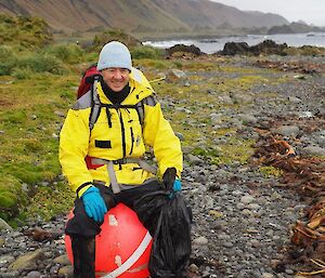 A man wearing a yellow jacket and beanie sits on a large pink buoy found washed up on the beach