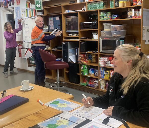 one person stands at a whiteboard, another at a radio console and a woman sits at a table in front of maps