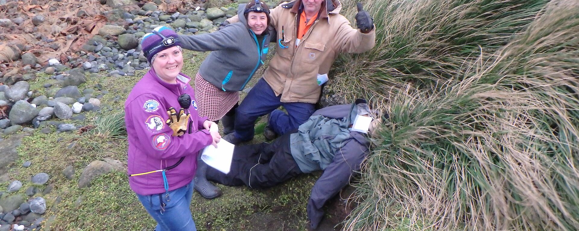 three people stand around a dummy laying in the tussock