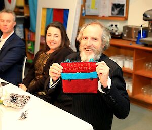 a man with a beard holds up a red embroidered bag saying Macca Midwinter 2019