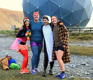 four women in fancy dress stand in front of a satellite dome