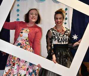 two women stand in party frocks holding a white photo frame in front of them