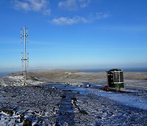 a small green tubular hut stands next to a satellite tower on top of a snow dusted plateau