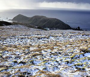 a view of a snow dusted plateau with a narrow view of an Isthmus and a research station with a headland and Southern ocean in the distance