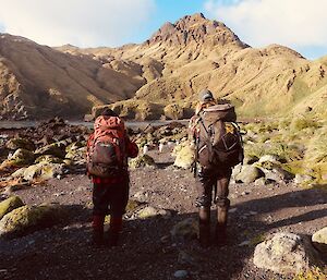 two women carrying bakpacks stand in front of a cove and mountain ridge above them. One points a walking stick into the distance.