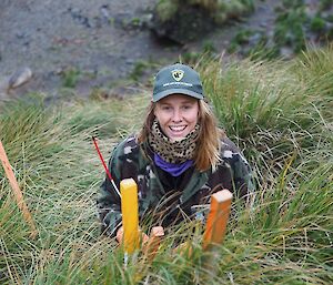 A woman wearing a cap sits in tussock grass with coloured wooden markers in front of her