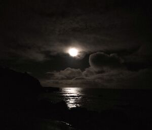 A bright full moon appears from behind dark clouds reflecting on the water