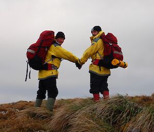 Two men wearing yellow jackets, backpacks are beanies are holding hands looking back at the camera from the crest of a hill.