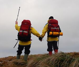 Two men in yellow jackets with backpacks stand with their backs to the camera. one holds a walking stick up in the air