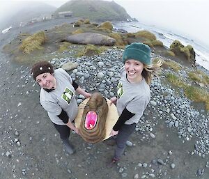 Two women hold an elephant seal cake outside with the station and an elephant seal in the background