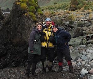 Two women and one man stand in front of a rocky outcrop on a beach