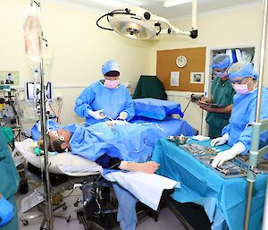 A patient lays on a surgical bed and a doctor simulates an appendectomy surrounded by three assistants in surgical scrubs