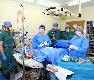 Five people in surgical scrubs and masks stand around a patient in a hospital bed