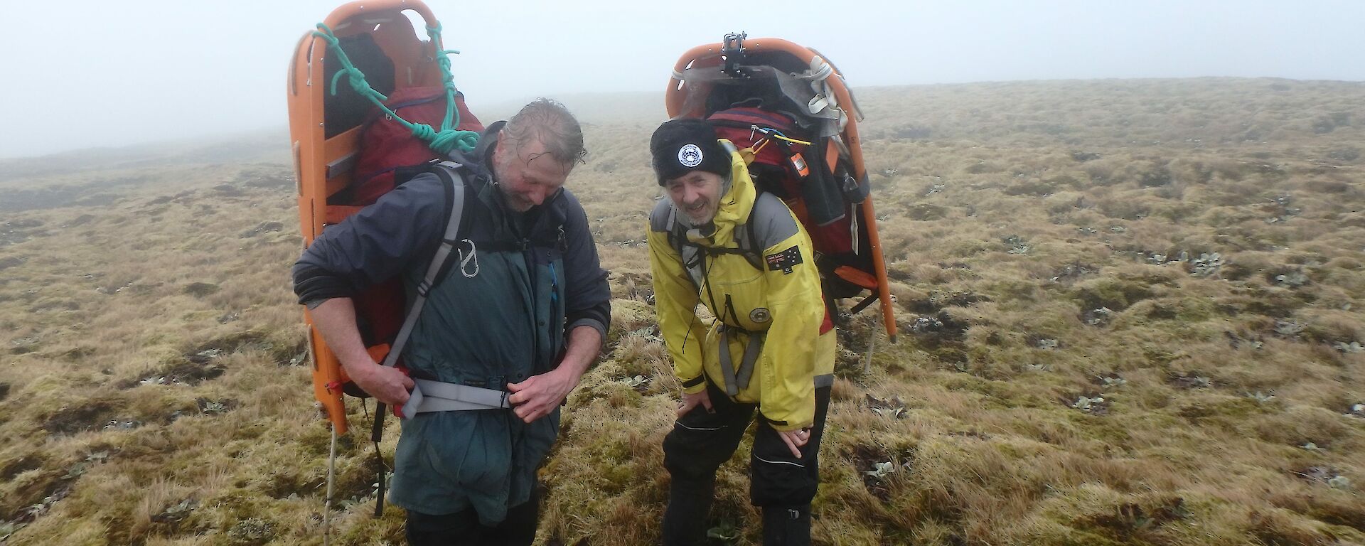 Two men with half a stretcher strapped to their backs stand on a grassy plateau
