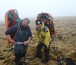 Two men with half a stretcher strapped to their backs stand on a grassy plateau