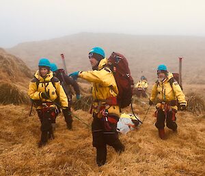 A man in a yellow jacket and blue helmet leads his team mates dragging a stretcher along the grass