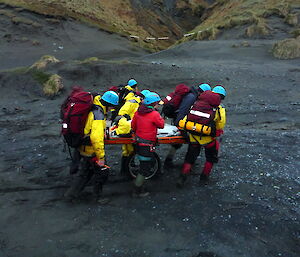 A team of people in rescue clothing carry a stretcher balancing on a rescue wheel along the beach