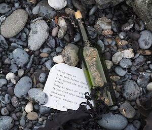 A green bottle lays on a pebbel beach with a typed note next to it