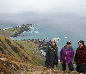three women stand on the edge of a steep cliff with the southern ocean behind them