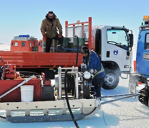 Two men pump fuel from a truck with fuel drums