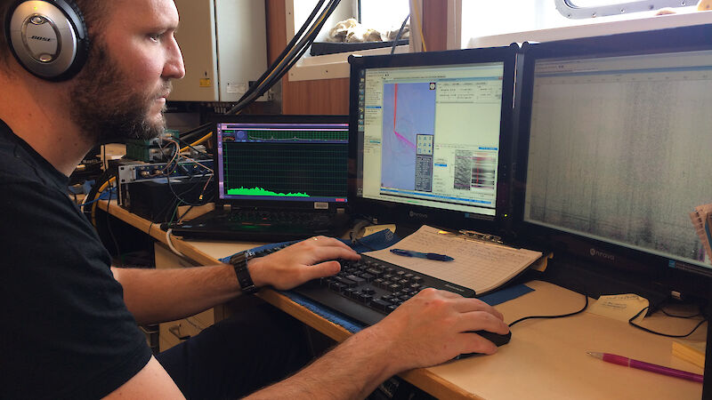 Dr Brian Miller listens for whales during a voyage, with a computer screen in front of him showing sounds.