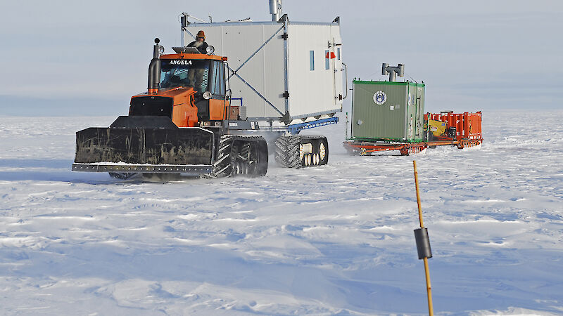 A tractor towing a shipping container on a sled across the Antarctic ice sheet.